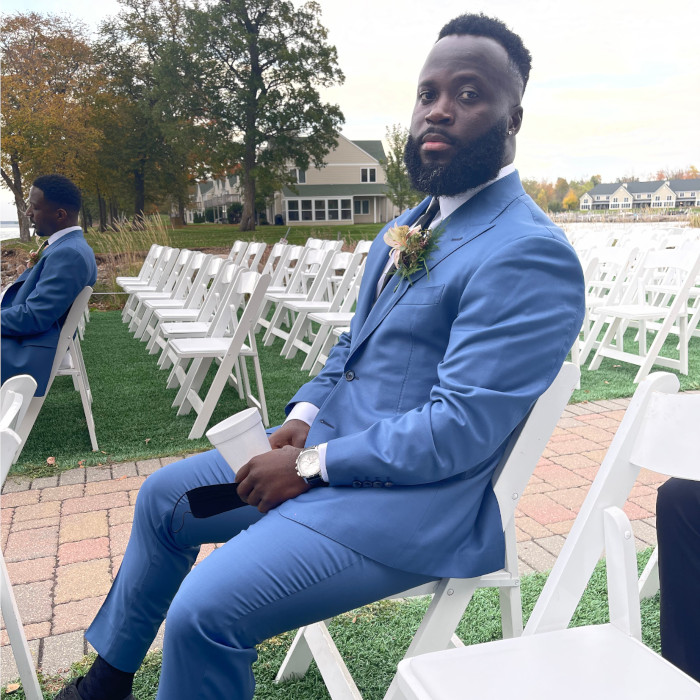 A man wearing a light blue suit is sitting in a row of white chairs.